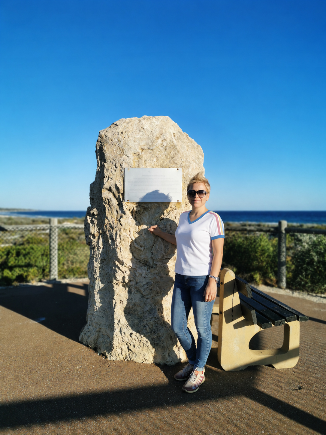 I wear my white V-neck T-shirt with blue bands and coloured shoulder stripes beside a stone monument by the sea.