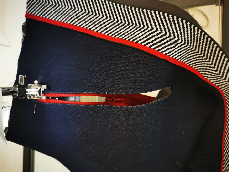 Presser foot beside an open red zipper, stitching over the existing line around the pocket opening.